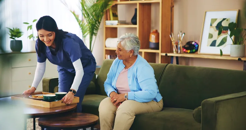 A woman in blue scrubs bringing a tray of tea to an older woman on a sofa