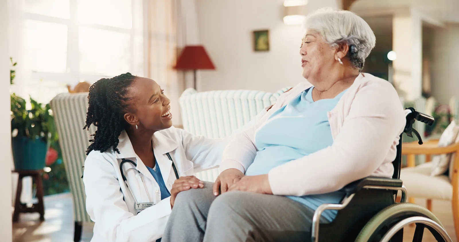 A smiling woman in a white coat with a stethoscope talking with an older woman who uses a wheelchair