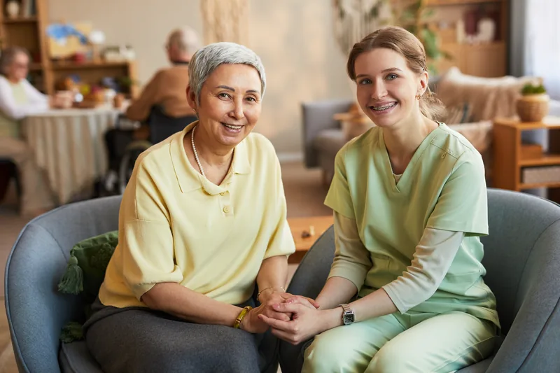 An older woman and a woman in pale green scrubs seated together holding hands