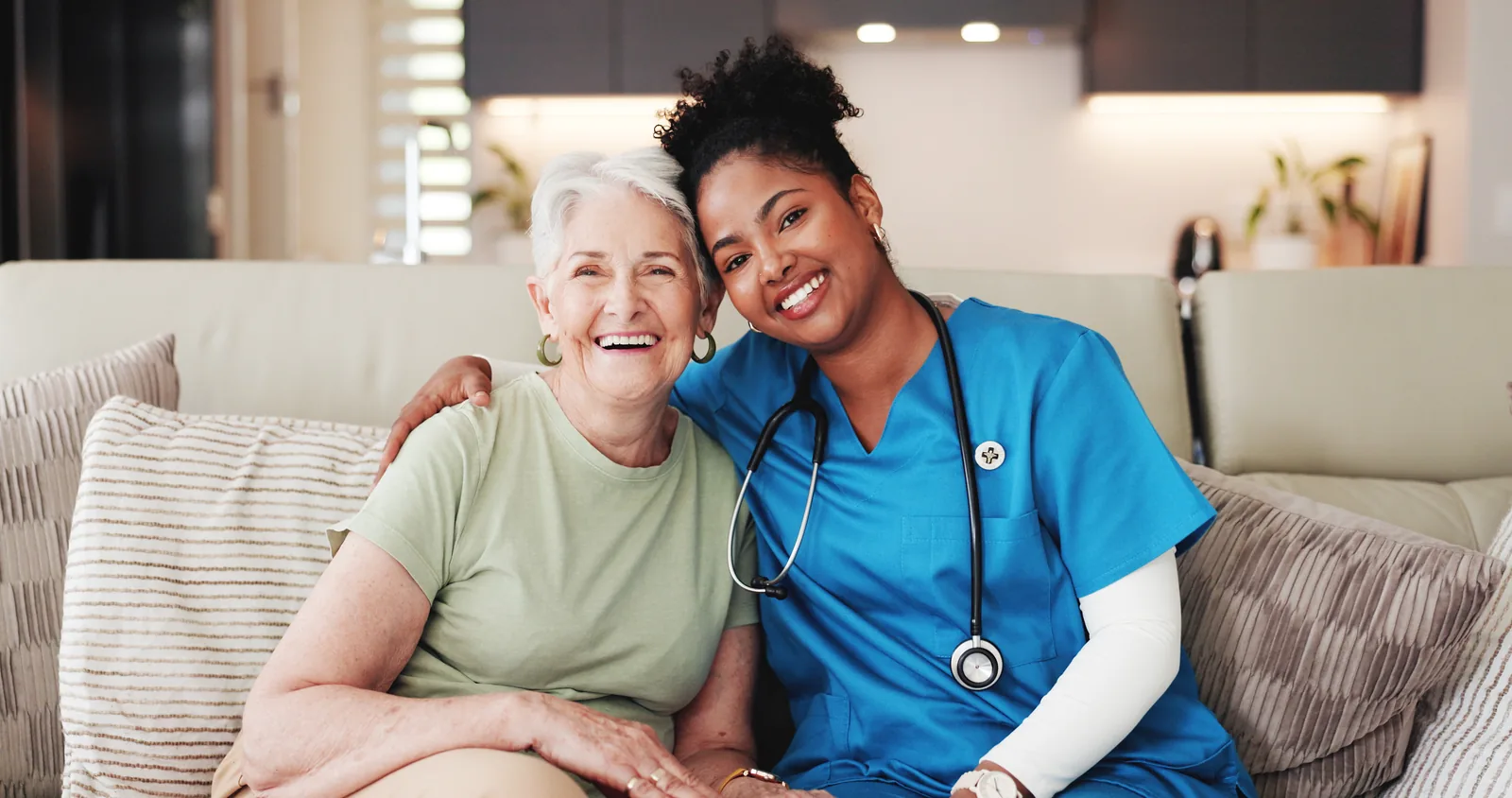 A woman in blue scrubs with a stethoscope sitting beside a smiling older woman on a sofa