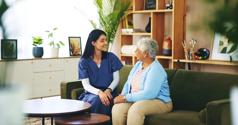 A woman in navy scrubs and an older woman talking together on a sofa