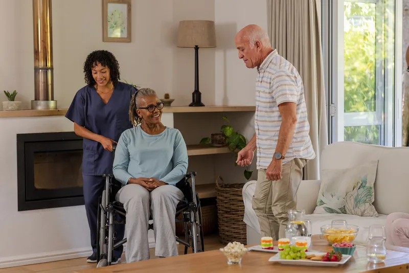 A woman in navy scrubs with an older woman in a wheelchair and an older man in a living room