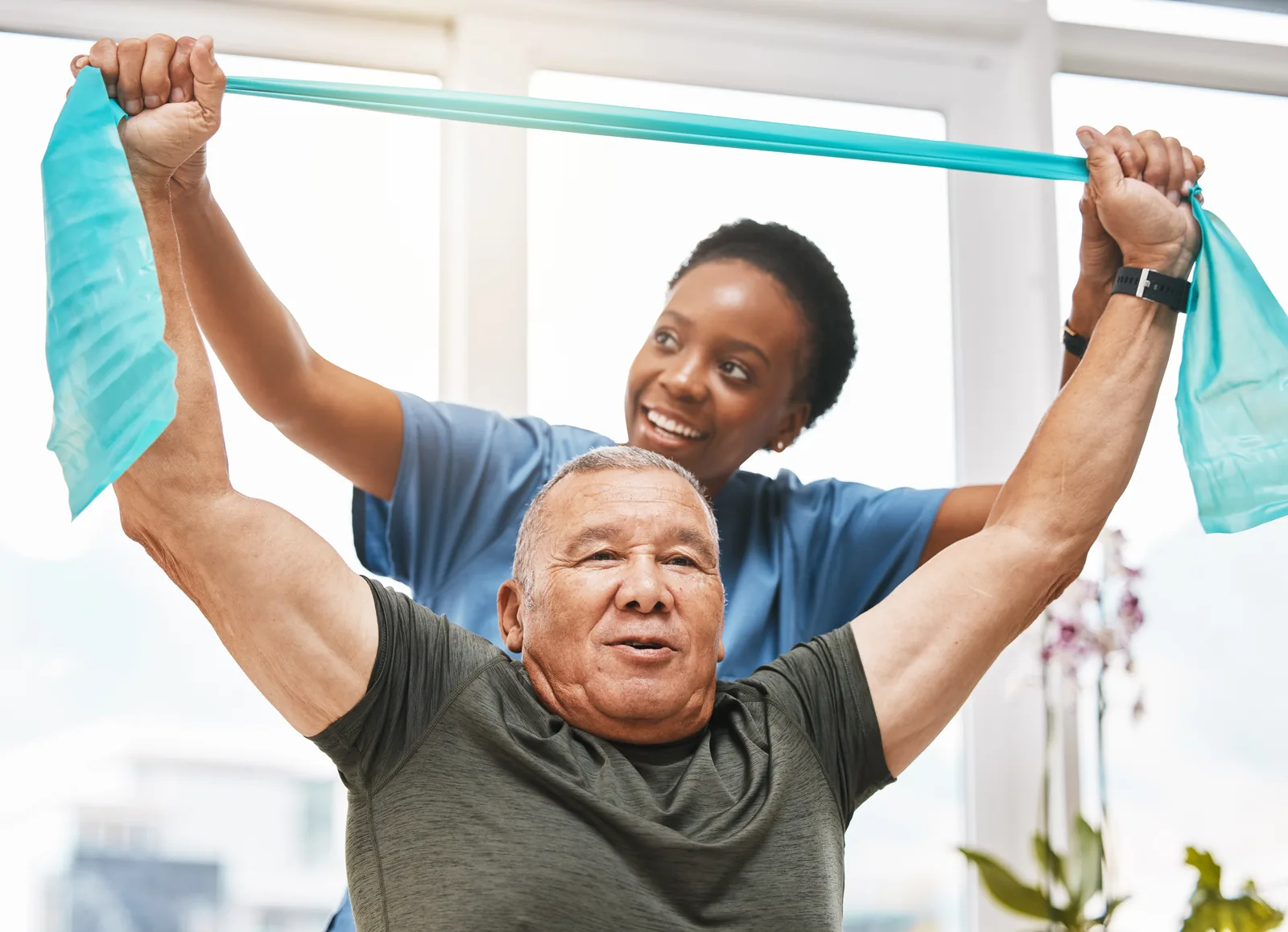 Older man raising a turquoise resistance band overhead with support from a woman in blue scrubs
