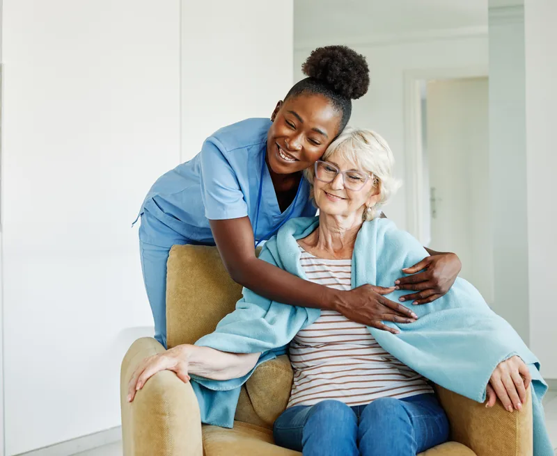 A woman in light blue scrubs hugging an older woman wrapped in a blanket