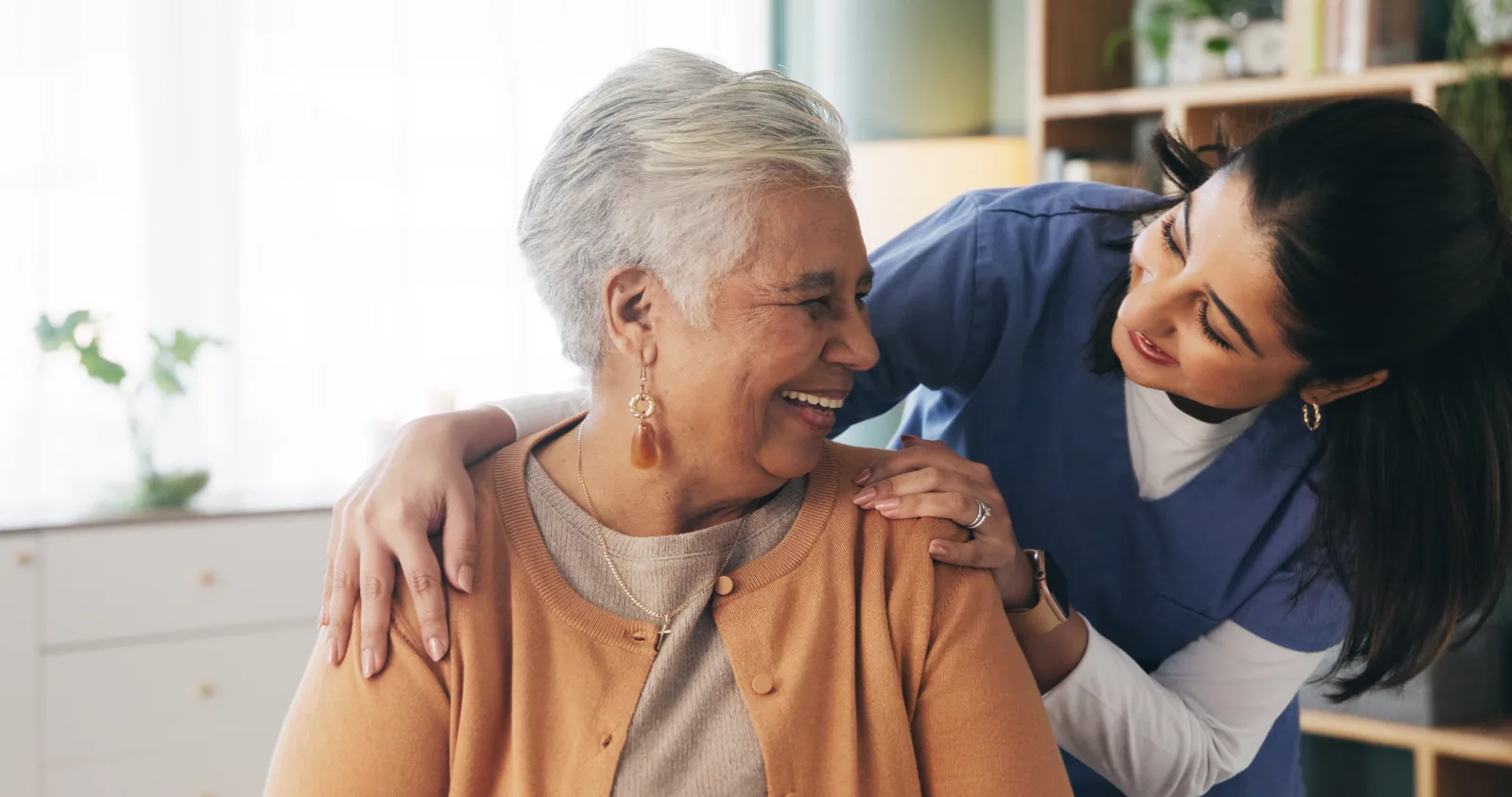 A woman in blue scrubs with her arm around a laughing older woman at home