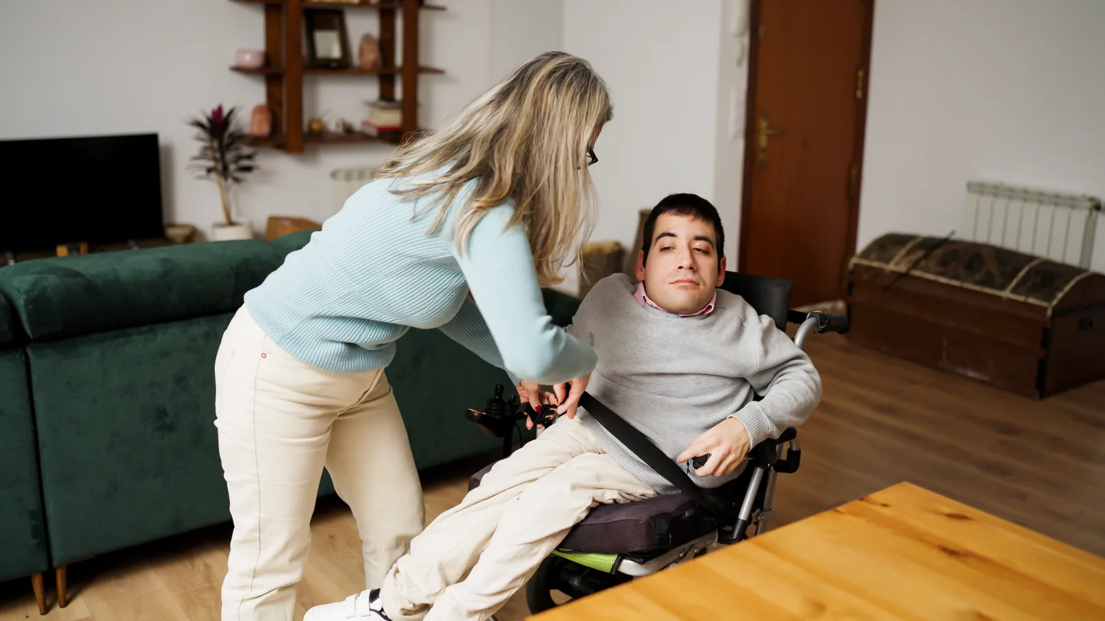 A woman assisting a man who uses a wheelchair in his living room