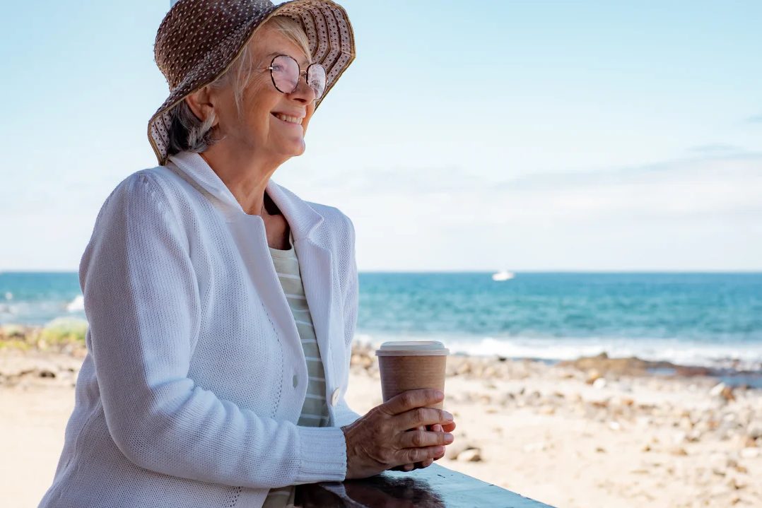Safety Tips for Seniors eqc Woman wearing heat, holding a cup of coffee by the side of a beach
