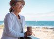 Woman wearing heat, holding a cup of coffee by the side of a beach