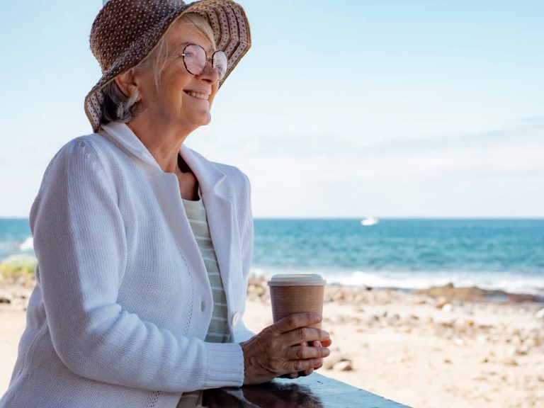Woman wearing heat, holding a cup of coffee by the side of a beach